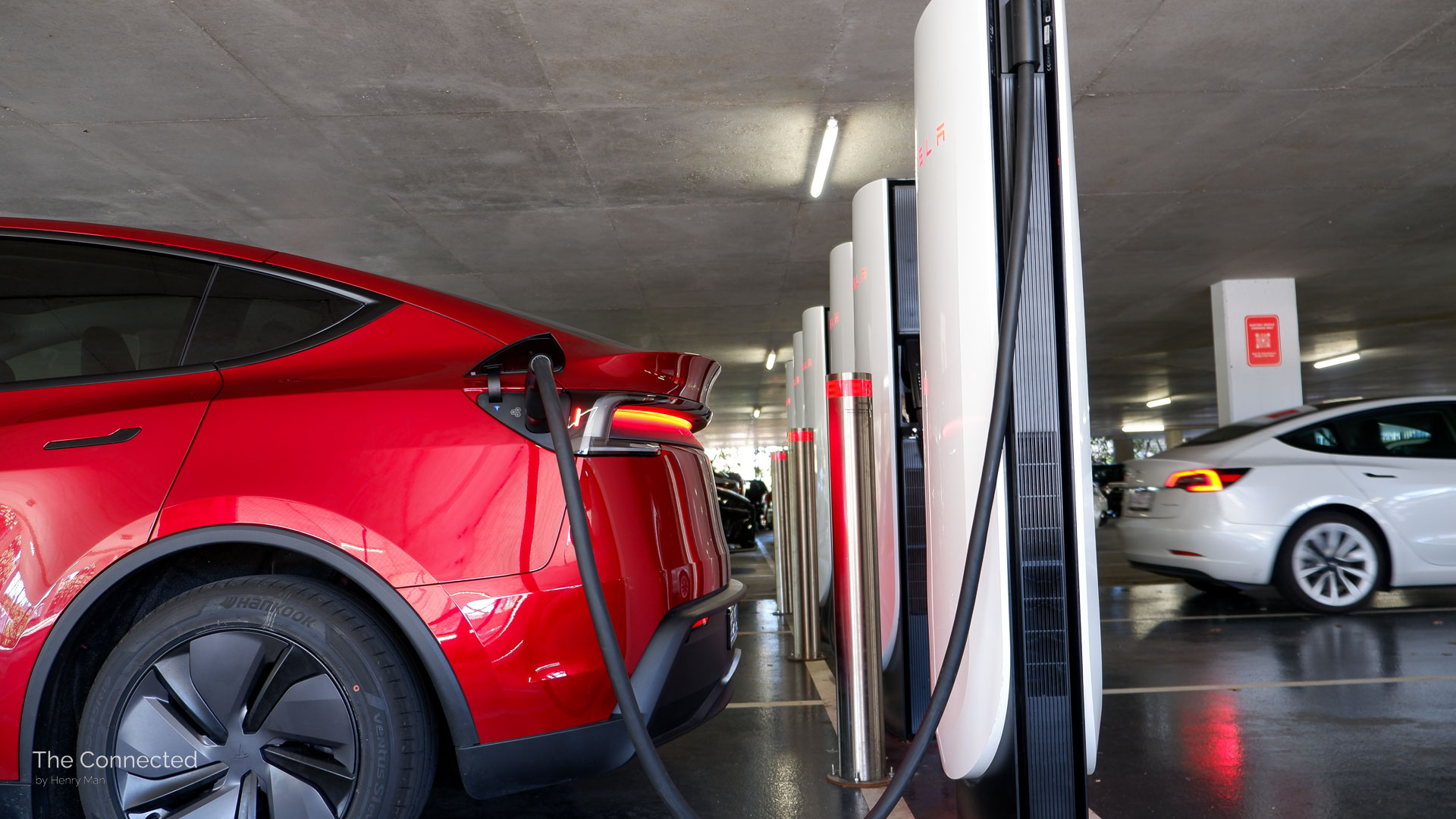 2026 Tesla Model Y RWD Ultra Red charging at V4 Supercharger with white Model 3 reversing in behind