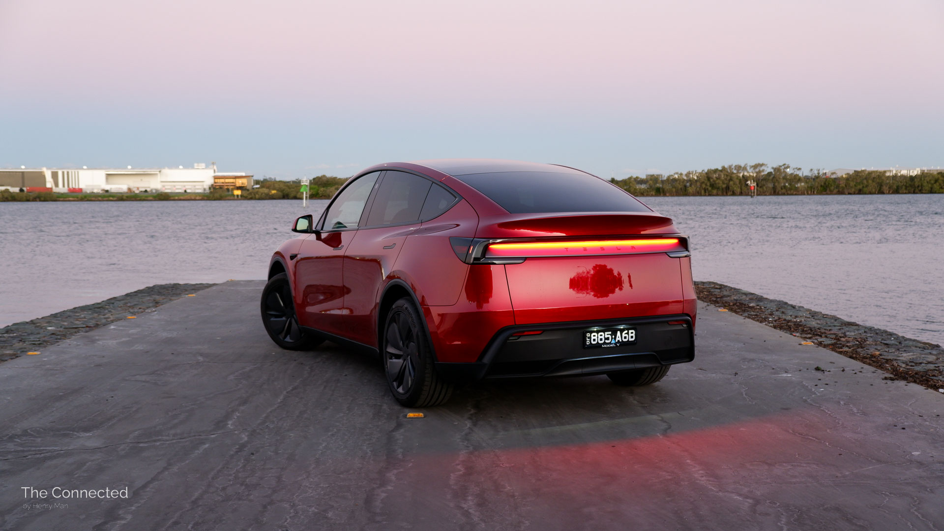 2026 Tesla Model Y RWD Ultra Red rear-quarter view with reflective light bar glowing onto the ground in the evening