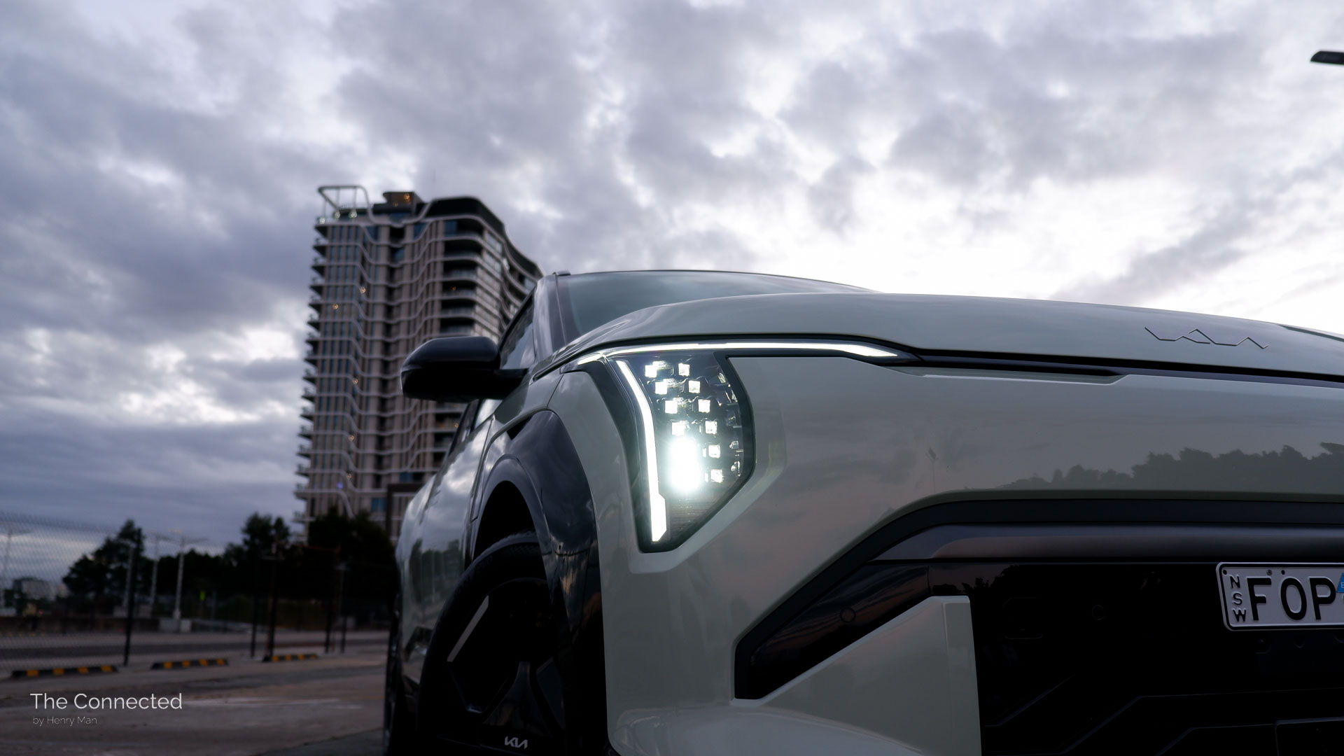 2026 Kia EV3 GT-Line front LED headlights with cloudy sky behind and city apartment