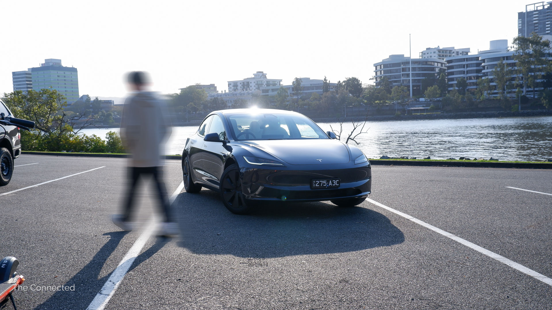 Man walks past 2025 Tesla Model 3 with the city behind in the afternoon