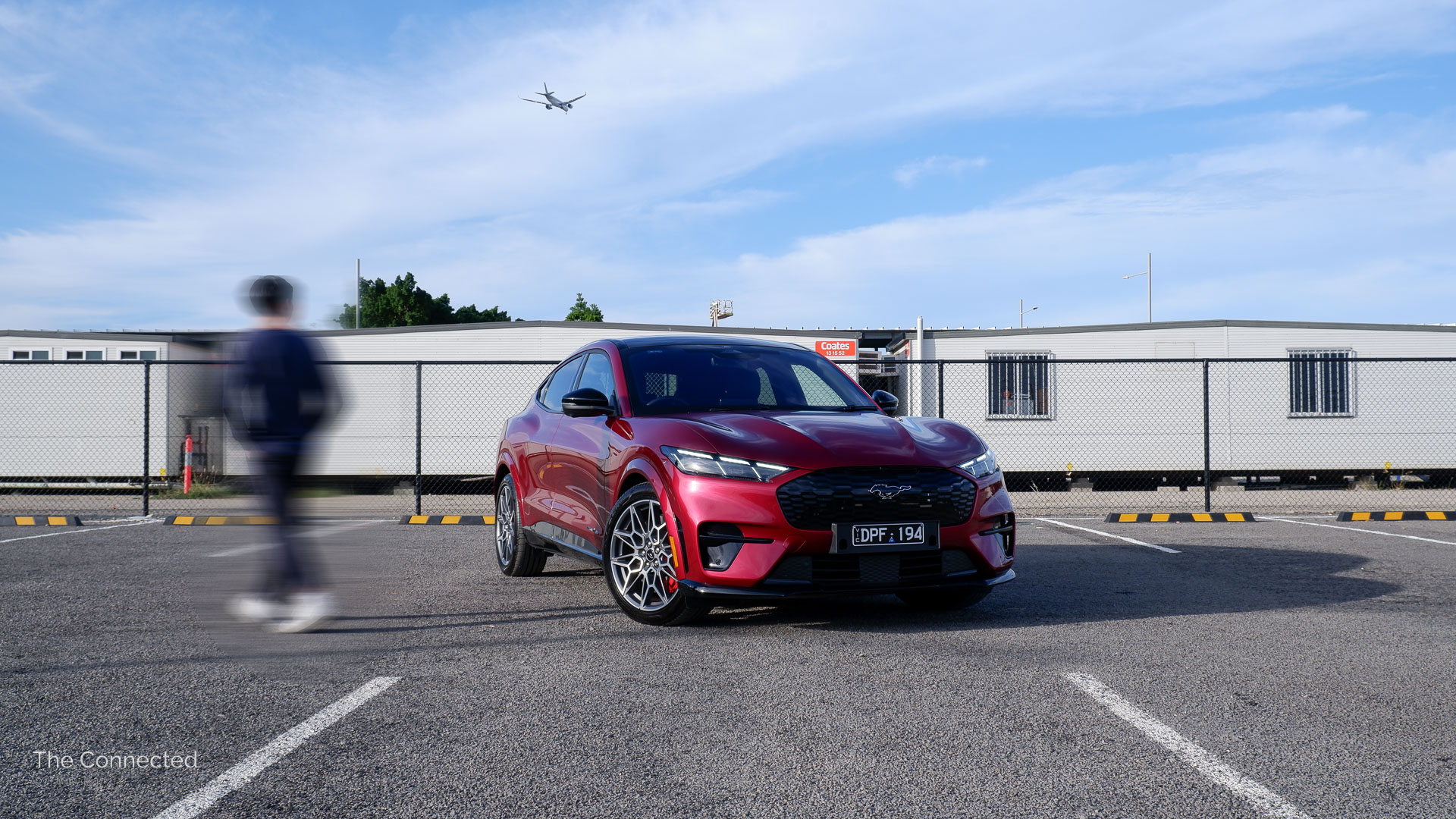 Man walks past the 2026 Ford Mustang Mach-E GT with an aeroplane in the sky