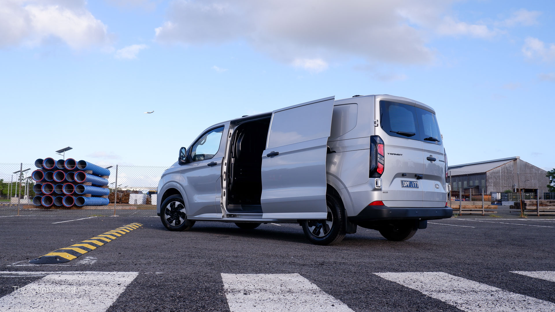 2026 Ford Transit Custom PHEV Trend rear-quarter view with sliding door open, blue pipes behind, and an aeroplane passing