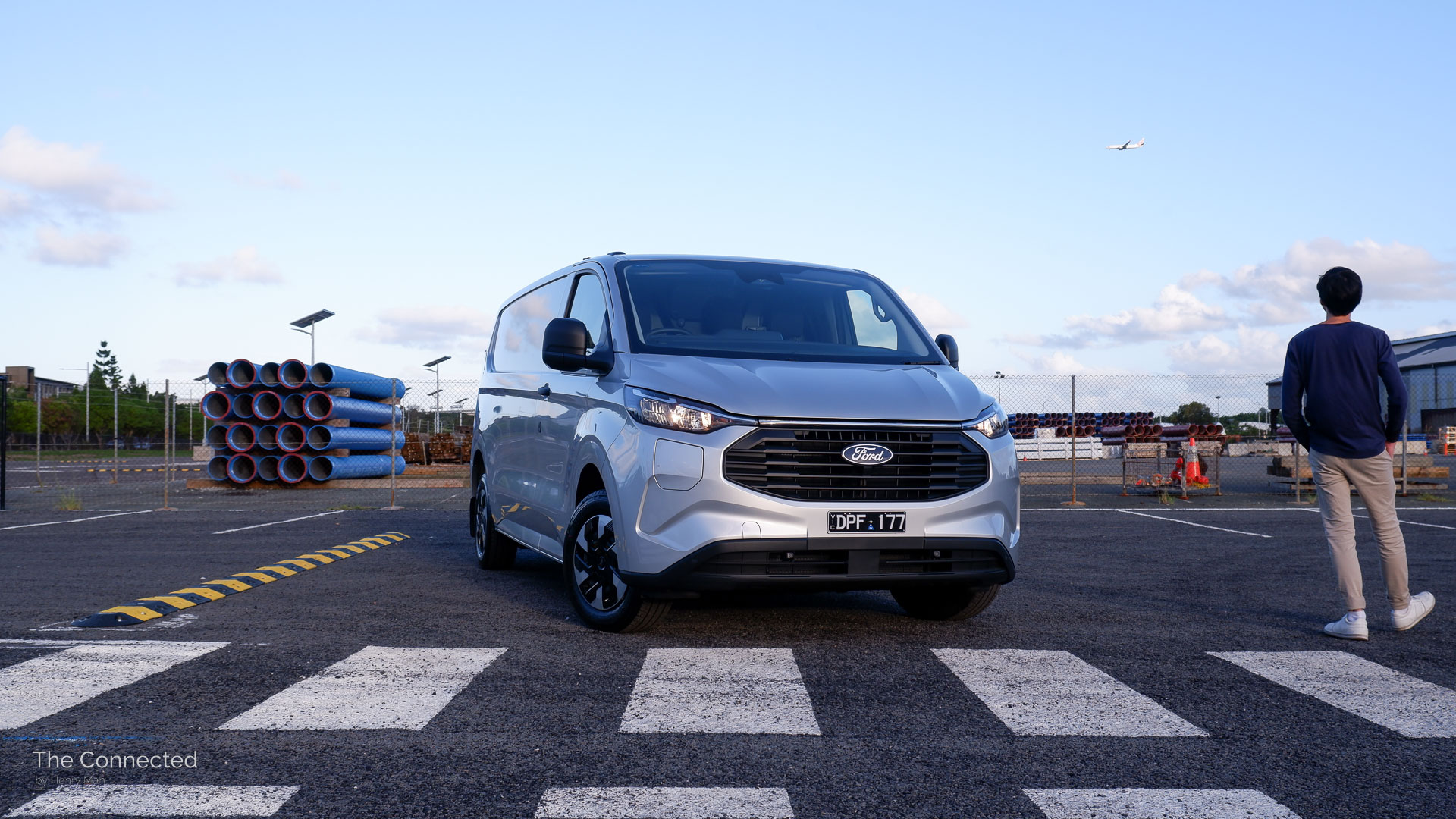 Man walks past the 2026 Ford Transit Custom PHEV Trend with an aeroplane passing in the sky and blue pipes behind