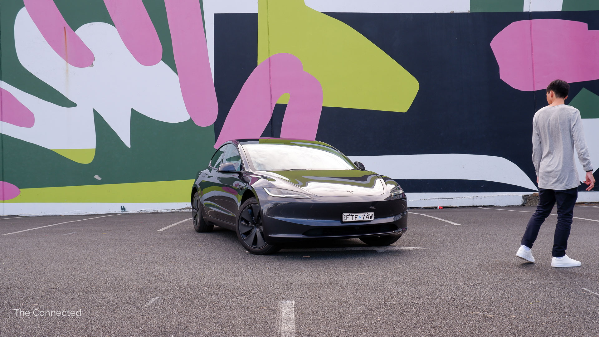 Man walks past the 2026 Tesla Model 3 Premium Long Range RWD with an artwork wall in the background