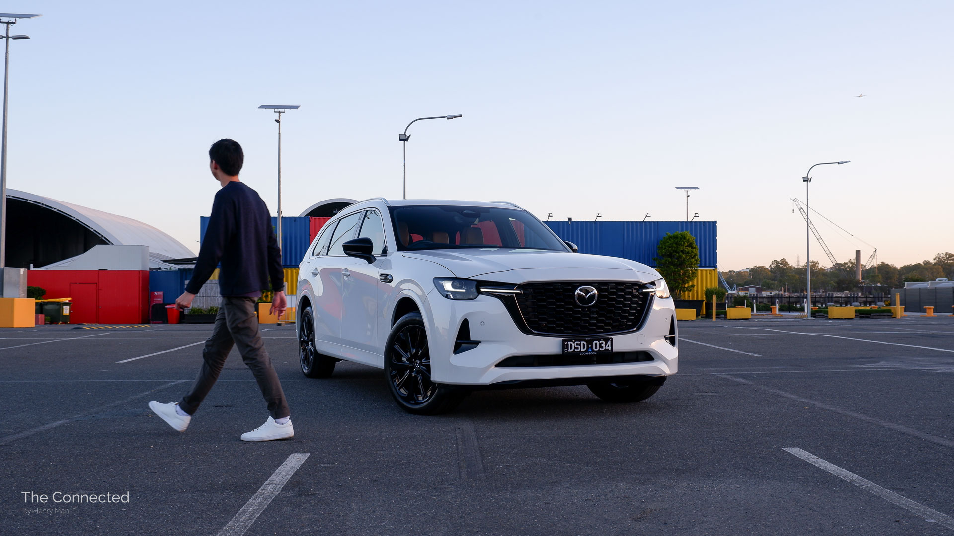 Man walks past the 2026 Mazda CX-80 P50e Azami SP during the sunset with colourful shipping containers behind