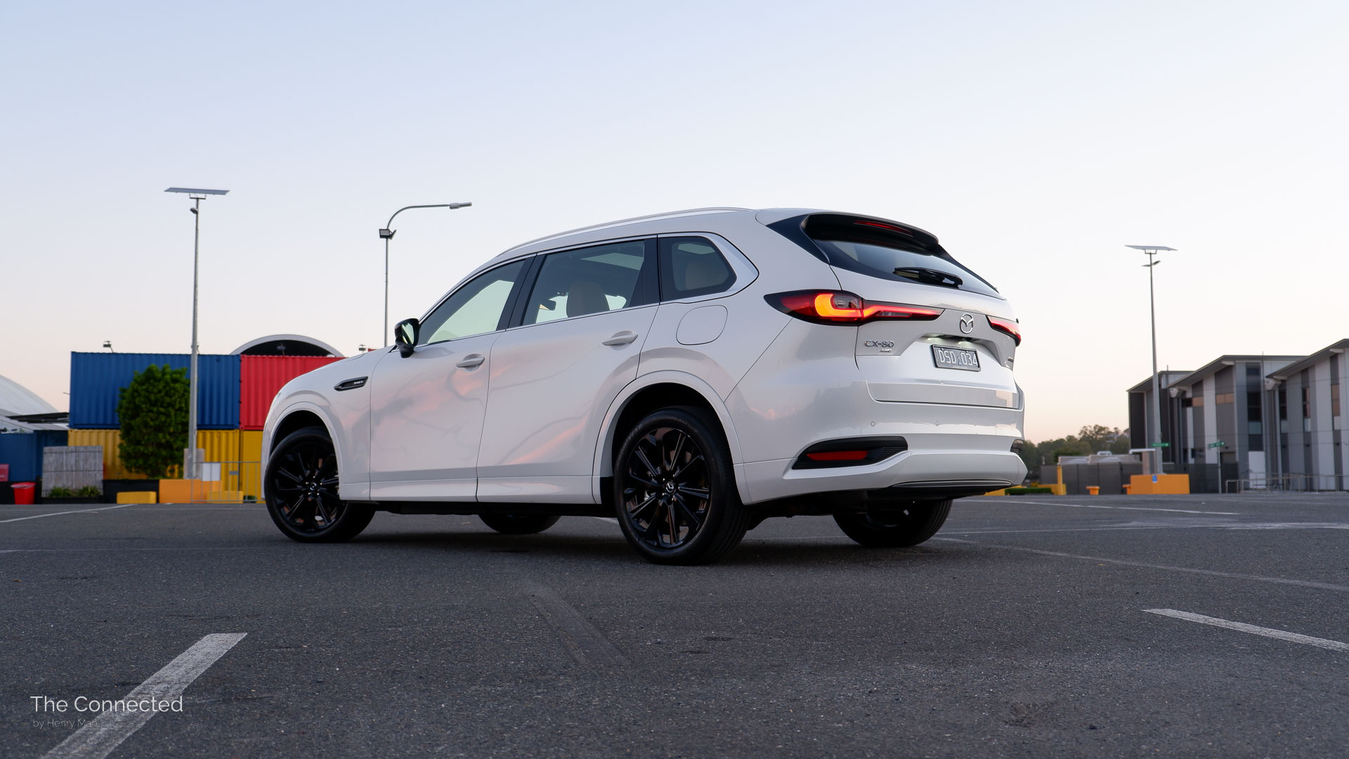 2026 Mazda CX-80 P50e Azami SP rear-quarter view during the sunset with colourful shipping containers behind