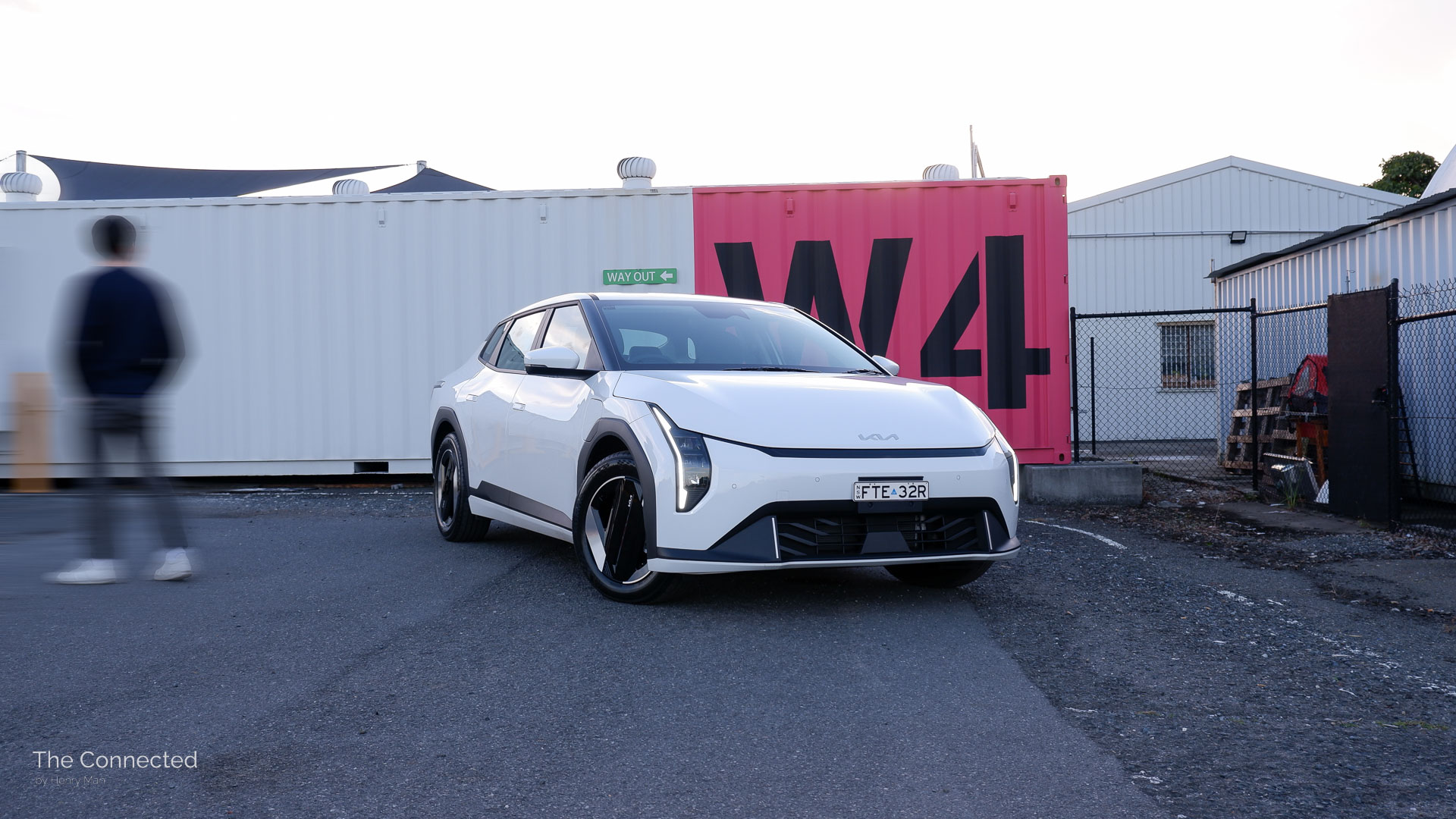 Man walks past the 2026 Kia EV4 Air  with a white shipping container behind painted with 'W4' in pink and black